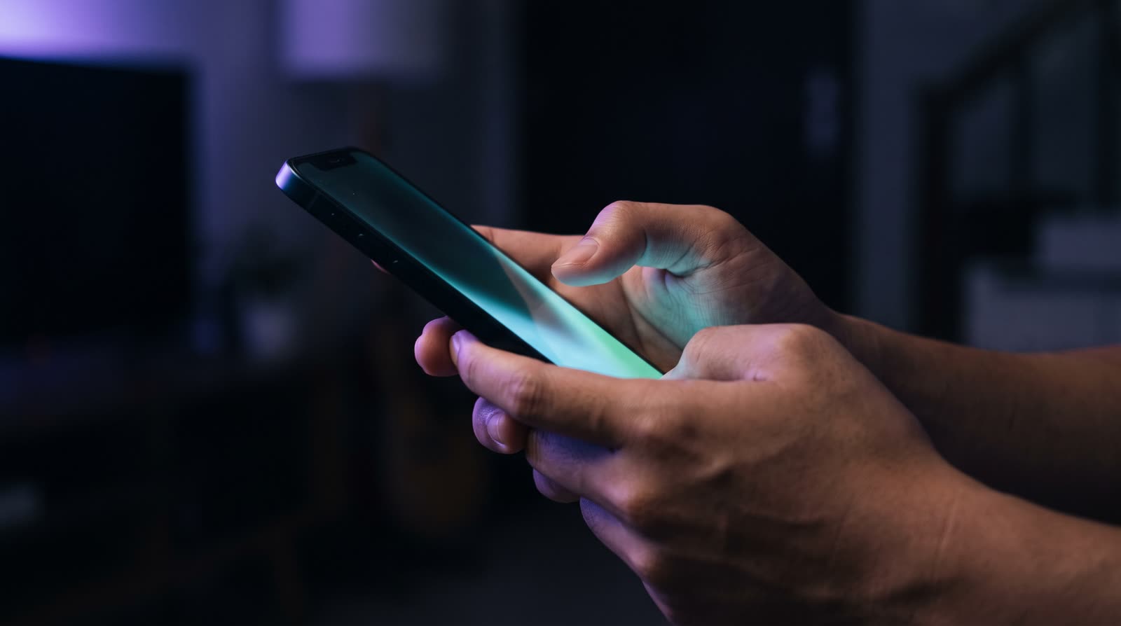 Tangan Indonesia memegang smartphone di ruang yang remang malam hari; layar ponsel menyala sian lembut, satu cahaya aksen ungu dari sudut kiri atas.