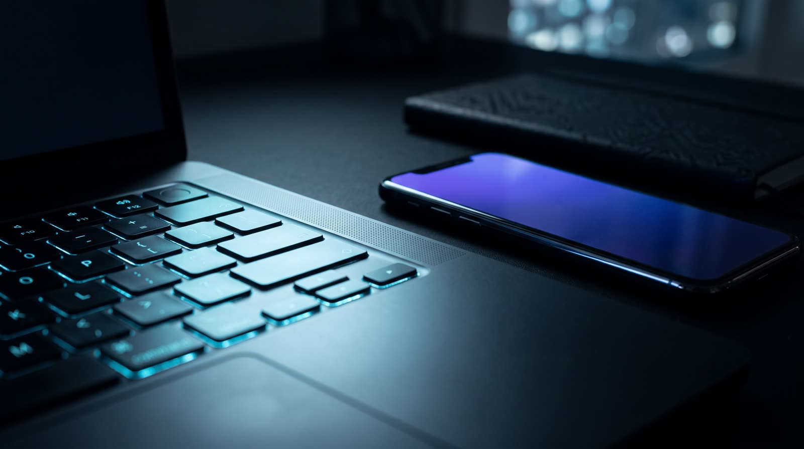 Makro meja kerja gelap malam hari: keyboard laptop ber-backlight sian, ponsel di sebelahnya menampilkan layar bergradasi violet-biru.