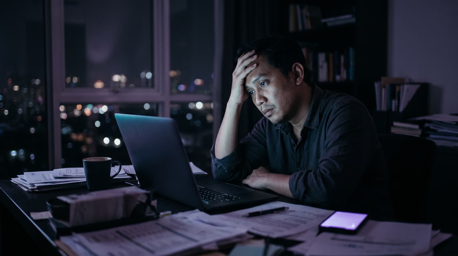 Pemilik usaha Indonesia sendirian di home office gelap pukul 2 pagi, menatap laptop yang menyala sian dan ponsel bercahaya violet, kertas berserakan di meja.