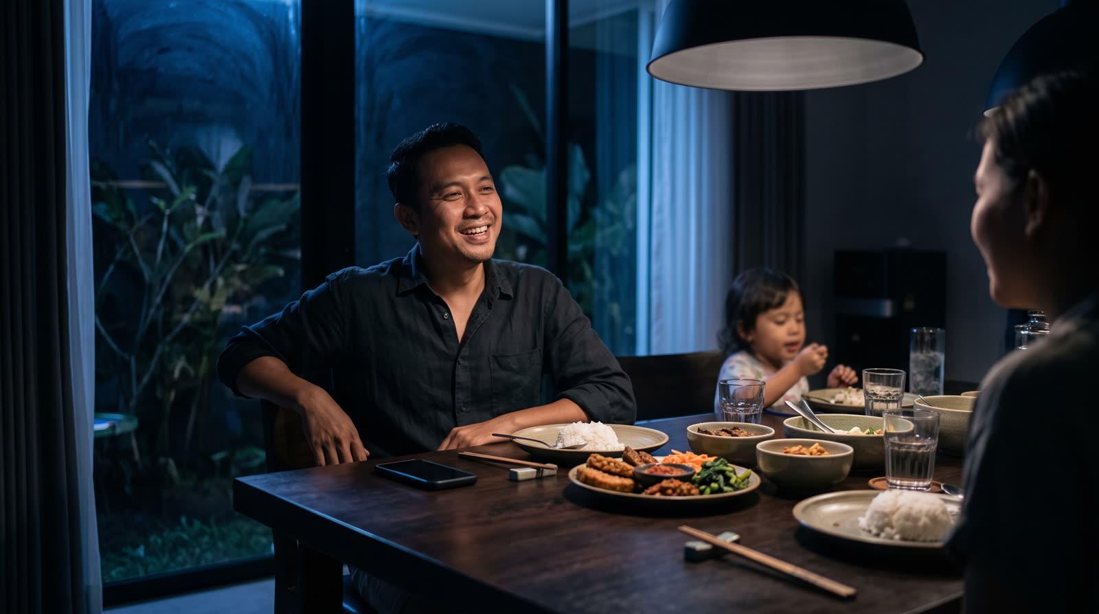 Keluarga Indonesia makan malam di rumah modern dengan cahaya pendant hangat; sang pemilik usaha duduk di kepala meja, ponsel diletakkan terbalik di sampingnya.